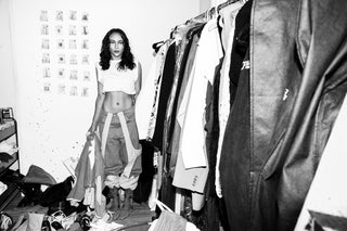 Black and white photo of a fashion model standing by a clothing rack while wearing jeans by designer Revive.